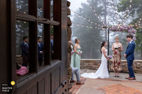 Groomsmen And The Wedding Party Are Reflected In A Window During The Ceremony At Historic Boettcher Mansion In Golden, Colorado. During the ceremony at Historic Boettcher Mansion in Golden, Colorado, groomsmen and the wedding party are artistically reflected in a window, adding a unique visual element to the celebration.
