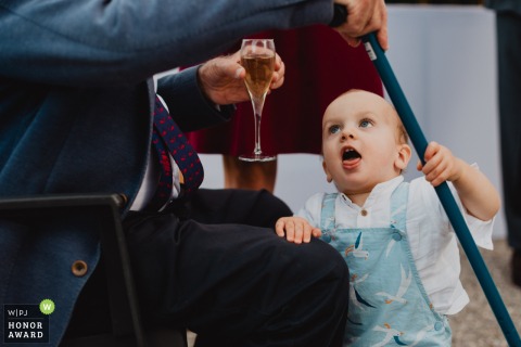 Grandfather Sips Champagne As Playful Grandson Clings To Leg During Outdoor Cocktail Hour At A Château In Meuse During the outdoor cocktail hour at a château in Meuse, a grandfather sits sipping champagne while his playful grandson clings to his leg, sticking out his tongue in delight.