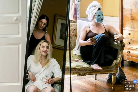 Relaxing With Masks And Champagne, Bride And Bridesmaid Prepare Together At Château De Montplaisant, Montagnat, Ain, France. Château de Montplaisant in Montagnat, Ain, sets the stage for bridal preparations, where a bridesmaid in a hydration mask and the bride with her hair getting done relax together with glasses of champagne.