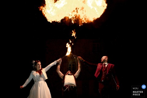 Evening At Domaine De La Chartrognière, Drôme, France, Sees The Bride Make A Face While Fire-Breathing Gasoline Is Sprayed. The evening at Domaine de la Chartrognière in Saint-Thomas-en-Royans, Drôme, features the newlyweds participating in a fire-breathing show, with the bride making a face as gasoline is sprayed on her.