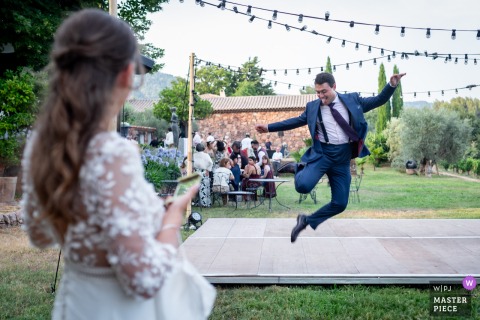   At Domaine de Grandpré in Puget-Ville, Var, France, the groom leaps with joy and heel clicks as he joins his wife on the dance platform, radiating happiness during the celebration.