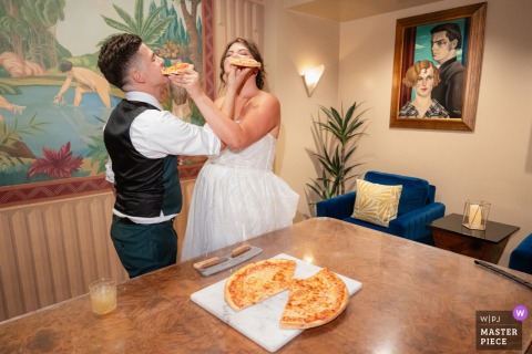  During the reception at The Oviatt in downtown LA, the bride and groom share a fun and unconventional touch as they cut their pizza together, surrounded by laughter and applause.