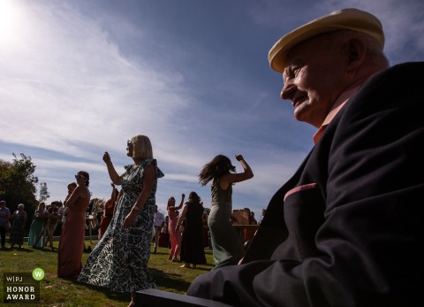 Outside the venue in Ille-et-Vilaine, Granddad enjoys the view of guests mingling and celebrating during cocktail hour, observing the lively party from a quiet corner at the wedding reception party.