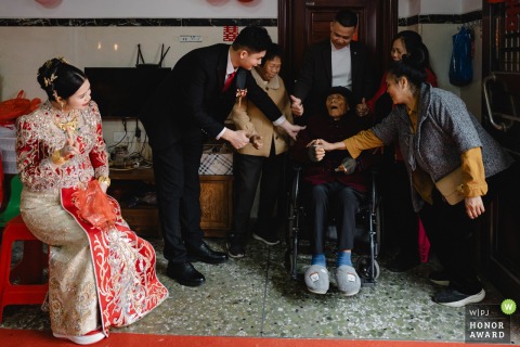 In Guangdong, four generations unite in the groom’s ancestral home as the groom’s parents and grandmother gently guide his great-grandfather for a meaningful family portrait, marking a treasured moment.