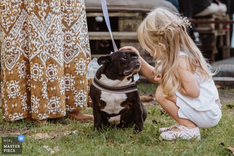 At Cascina Capitanio In Novara, Italy, Bride’s Niece Shares A Touching Moment With The Family Dog During The Wedding. At Cascina Capitanio in Novara, Italy, the bride’s niece crouches to meet the family dog eye-to-eye, sharing an affectionate connection during this special family celebration.