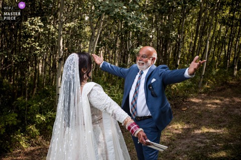   Beaming with happiness, the bride and her dad walk toward each other with open arms, ready for a warm embrace upon arrival at the wedding ceremony location in the Alberta woods..