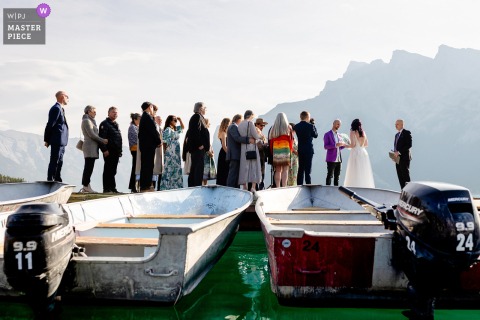 Alberta's Majestic Mountains Frame Wedding Vows On A Lake Dock, Creating A Stunning And Memorable Ceremony Moment. The ceremony takes place on a lake dock with mountains as the backdrop, giving the couple’s vows a breathtaking setting for their special moment in Alberta.