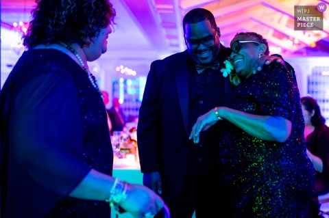   Laughter fills the air at Woodland Park, NJ, as the groom enjoys a lighthearted moment with family members, surrounded by good spirits and blue/purple lights.