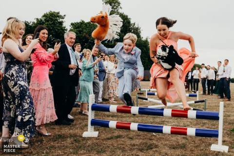 Young Guests Find Pure Delight Racing Hobby Horses At Worcester Lodge, Badminton, Filling The Celebration With Playfulness. Young guests at Worcester Lodge in Badminton find delight in a lighthearted round of hobby horse racing, their laughter and playful spirit energizing the outdoor wedding celebration.
