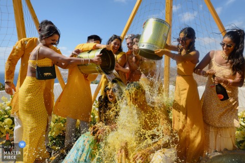 Friends In Mykonos, Greece Surprise Bride And Groom With A Bucket Of Cold Water, Sparking Laughter On The Big Day. At Royal Miconian in Mykonos, Greece, friends surprise the bride and groom by dumping a whole bucket of chilly water on them, eliciting laughter and shock on their special day.