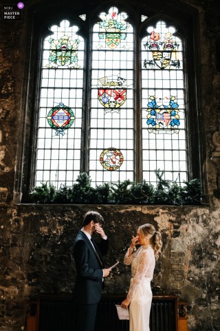   The bride and groom gently wipe away tears during their vows, emotion filling the ceremony location as they exchange heartfelt promises in front of the tall stained glass windows.