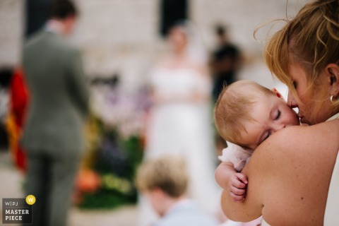   During the ceremony, a bridesmaid lovingly kisses her baby daughter, sharing a heartfelt moment between generations while guests watch at the ceremony location.