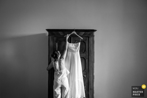   In a quiet room at Château De Saint-Martory, France, a black and white photograph shows a woman in pajamas reaching toward a bridal gown hanging inside an antique wardrobe.