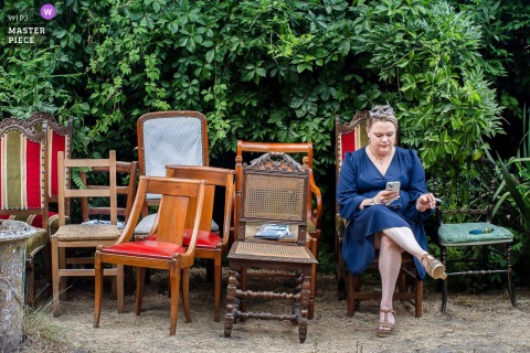In Largentiere, France, A Woman Sits Among Old Chairs, Creating A Nostalgic Scene In The Elegant Wedding Setting. In Largentiere, France, a woman sits alone outside surrounded by well-worn old chairs, adding character and a touch of nostalgia to the elegant wedding setting.