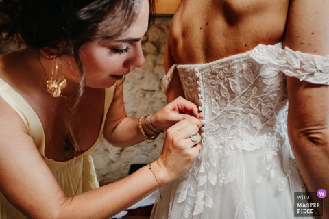 Friends Carefully Fasten The Bride’s Dress At Domaine Des Monts Du Lyonnais In Rhône, Preparing Her For Ceremony. Nimble fingers are required to button up the wedding dress at Domaine des Monts du Lyonnais in Rhône, as friends carefully help the bride prepare for her big moment.