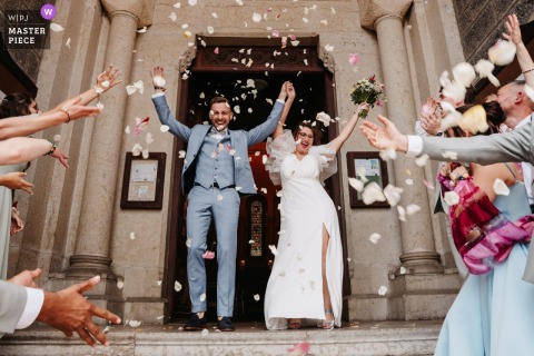   A triumphant exit at the church in Vaugneray, Rhône, sees the newlyweds showered with flower petals as they step onto the church steps, smiling and waving at their cheering guests.