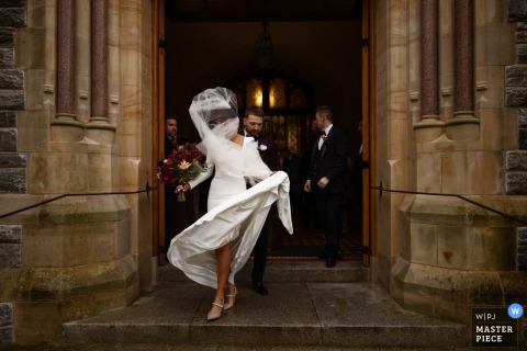   As she leaves Strabane Church in Northern Ireland, the bride’s veil and dress are lifted by a playful gust of wind, adding a touch of spontaneity to her exit.