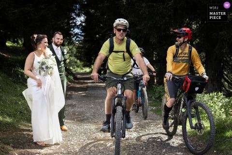 Couples Walk Hand-In-Hand Through Chatel, Haute-Savoie As Passing Cyclists Join The Scene On Their Wedding Day. The couple meander hand-in-hand through Chatel, Haute-Savoie as cyclists are passing through together on their journey.