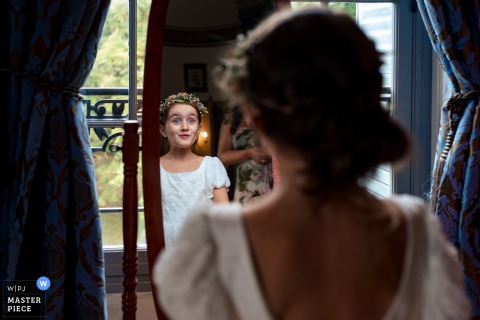   Eyes wide with wonder, a little girl admires her reflection in an ornate mirror at Chateau de la Canière, France, soaking in the fun of the wedding setting.