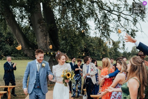 Confetti Fills The Air While Newlyweds Joyfully Walk The Aisle At Swanley Meadow In Cheshire, United Kingdom. Confetti fills the air as the bride and groom make their joyful walk down the aisle at Swanley Meadow in Cheshire, UK, surrounded by friends and family.