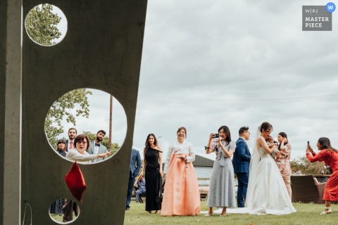 Bride Balances Family And Joy, Holding A Baby While Guests Play Beanbag At Pheasantry Brewery, Nottinghamshire, UK. While guests play a beanbag game at Pheasantry Brewery in Nottinghamshire, UK, the bride stands smiling and holding a baby, balancing joy and family during this lively celebration.