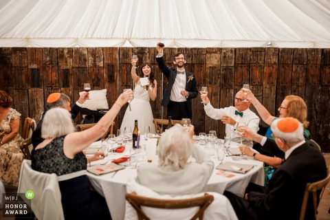 During wedding speeches at Burningfold Barn in Godalming, the couple proudly raises their glasses of wine in a joyful toast, celebrating with friends and family.