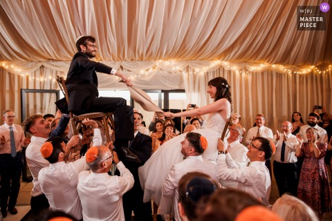 Cheers And Laughter Fill Burningfold Barn In Godalming, UK As The Couple Is Lifted On Chairs For Hora Dance. At Burningfold Barn in Godalming, UK, the bride and groom are joyfully lifted on chairs during the wedding hora dance, surrounded by cheering guests and lively celebration.