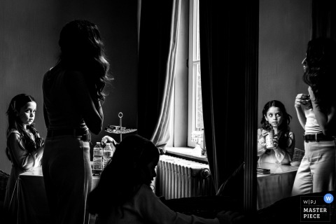 In A Wallonie Hotel Room, Soft Window Light Illuminates The Bride’s Daughter Adoringly Watching Her Mother Prepare. In a hotel room in Wallonie, the bride’s daughter gazes admiringly at her mother, with beautiful natural window light illuminating their quiet and intimate moment together.