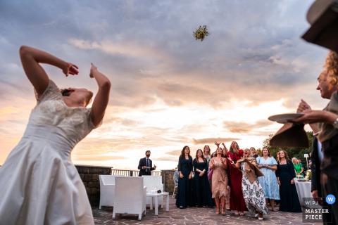   At sunset in Trieste, Italy, the bride tosses her bouquet as guests gather to catch it, the warm light adding atmosphere to the cherished tradition.