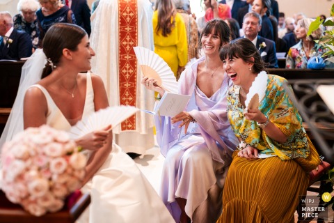   In Gorizia, Italy, two women guests laugh as a funny moment unfolds during the wedding ceremony, lightening the atmosphere and bringing smiles to faces in this beautiful Italian location.