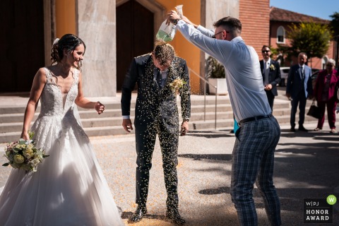 At a wedding in Udine, Italy, a friend gleefully empties a bag of rice over the groom’s head, celebrating the couple with a joyful and traditional gesture.