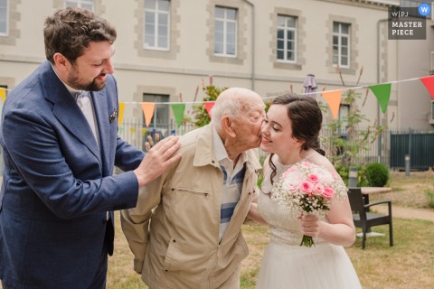 Tender Grandfatherly Affection Moves The Bride During A Family Moment At The Brittany Reception Venue In French Countryside. At the reception venue in Brittany, a tender kiss from her grandfather moves the bride, marking a sweet and memorable family moment during the celebration in this French region.