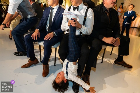   During the reception at NOOR Events in Pasadena, California, a child laughs as his father playfully holds him upside down during a lively wedding game, entertaining guests with their antics.