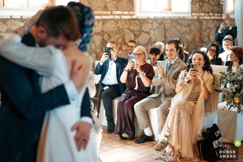 During a wedding at Borgo Colle Mentuccia in Roma, guests enthusiastically use their smartphones to capture the newlyweds lovingly embracing each other.