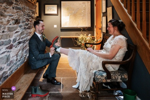   At Hause Hall Farm in Cumbria, England, the groom helps his new wife remove her boots on their wedding day, illustrating the couple’s comfort and caring in a rustic setting.