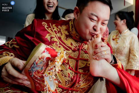   At home during the wedding reception in Shanxi, friends playfully coax the groom into kissing the bride’s feet, sharing laughter and lively tradition as guests watch and cheer them on.