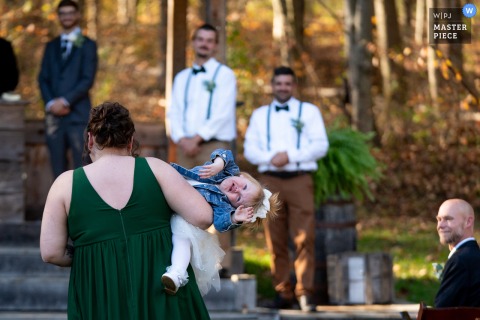   At Hinckston Run Farm in Johnstown, PA, a bridesmaid carries a crying flower girl down the aisle while the father of the groom watches the emotional moment unfold.