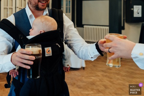 Through Gas Street Church in Birmingham, UK, a guest carries both beer and a baby, and the scene folds adult celebration and parental duty into one comic, exhausted frame.