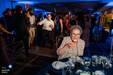 At Old Ranch Country Club in Seal Beach, California, the groom’s grandmother enjoys cake while the dance floor swirls beyond her, a sweet study in age, appetite, and timing.