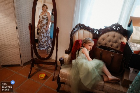 At Leo Carrillo Ranch in Carlsbad, California, a flower girl rests on a couch while the bride dresses nearby, an unguarded pause balancing ceremony preparation with childhood ease.