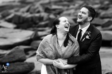 A couple in their wedding attire sharing a joyful laugh as they pose for portraits at the rocky shoreline of Acadia National Park, Maine