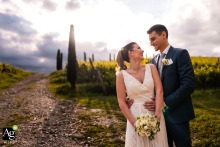 A happy bride and groom stand near a vineyard at sunset in Bosco Romagno, Cividale del Friuli, Italy. They smile and hold hands, surrounded by beautiful grapevines and a golden sky.