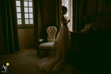 At Domaine des Gaillardoux in Lot, France, the bride stands silhouetted by window light during her preparations, creating a dramatic and elegant getting-ready image.