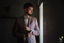 During preparations in Moselle, an elegantly dressed man in a pale pink suit and floral bowtie adjusts his watch by a softly lit window, his thoughtful expression enhanced by intimate, gentle light.