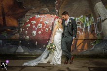 At The Old Fruitmarket in Glasgow, the bride and groom pose in front of a vibrant mural wall. This setting captures the lively artistic spirit of the city and their joyful celebration.
