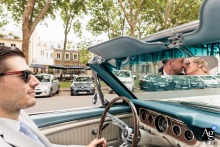 Bride and groom kissing in the reflection of a convertible car's mirror in Versailles, Yvelines, as the driver focuses on the city roadway.