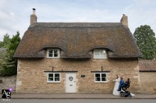 The bride and groom are photographed together in Northamptonshire, East Midlands, walking in front of a traditional thatched cottage with the groom seated in his wheelchair.