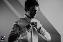 An artistic, soft-lit black-and-white portrait taken in a Budapest, Hungary, hotel room focuses on a thoughtful groom buttoning his shirt with eyes closed in calm anticipation of his wedding.