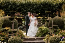 Romantic Symmetrical Wedding Portrait With Warm Light At Chenies Manor Buckinghamshire United Kingdom Chenies Manor, Buckinghamshire, United Kingdom — The bride and groom stand together in the gardens of Chenies Manor, captured in symmetrical framing with warm light highlighting their loving connection in this romantic wedding portrait.
