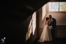 A wedding couple is artfully framed in a corner of Ebbergkirche Hemer, Germany, with vibrant colors from the stained glass windows casting a beautiful, painted glow on the adjacent stone wall.